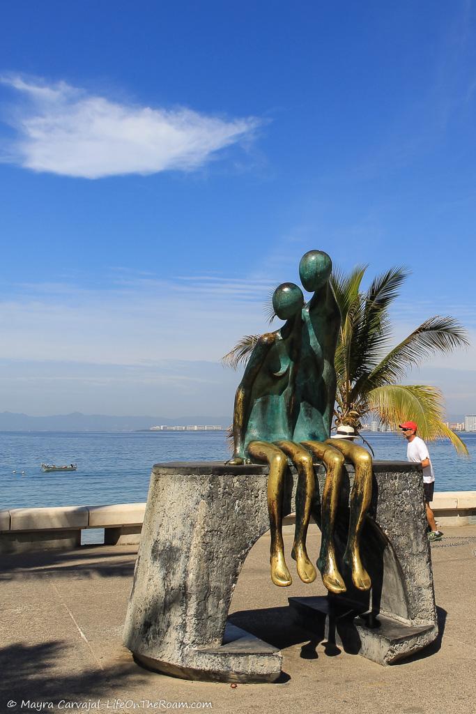 A bronze sculpture of two anthropomorphic figures seating side by side with the sea in the background s