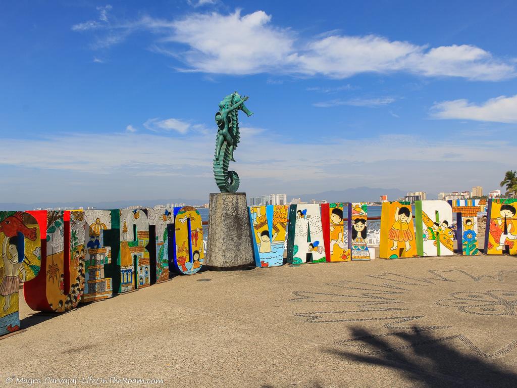 A sign with the Puerto Vallarta letters and a sculpture of a boy on a seahorse, on a seaside boardwalk