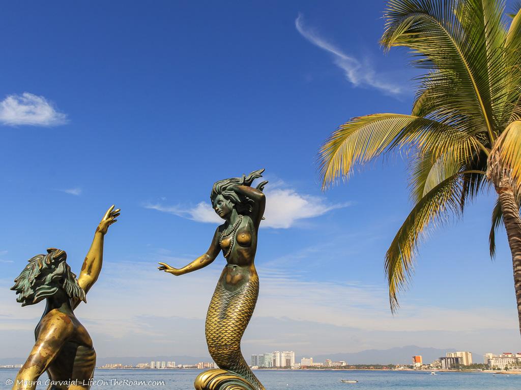 A bronze sculpture with mythological marine figures of a Triton and a siren, with the sea in the background