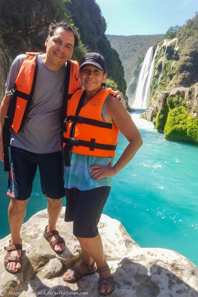 A couple standing on a big rock with a waterfall in the distance