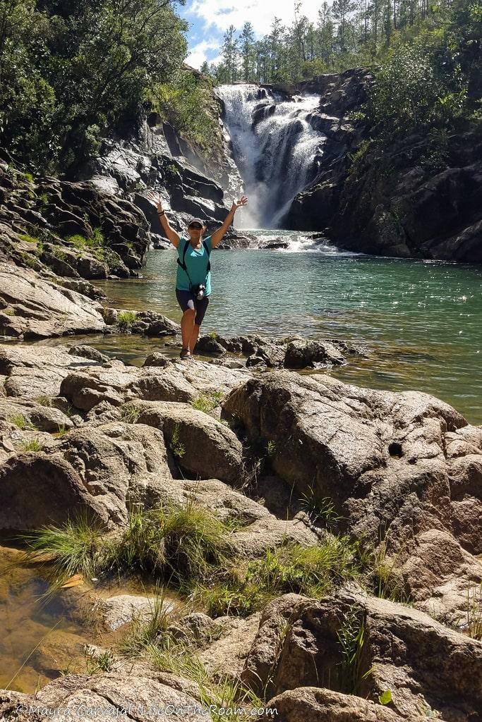 A woman standing on rocks next to a creek with a waterfall in the background