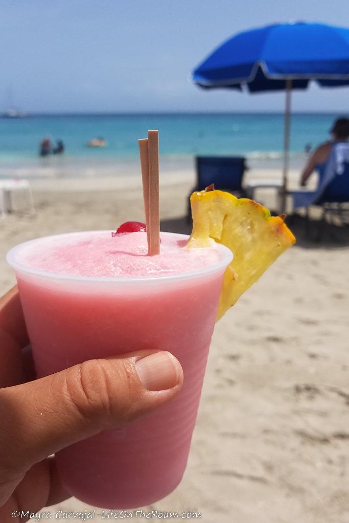 A hand holding a pink drink on a beach
