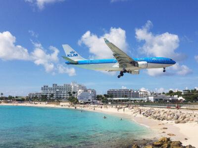 A large plane flying low over a beach with turquoise waters right before landing