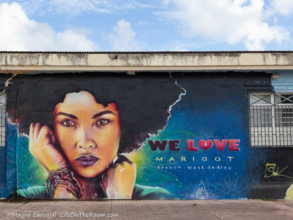 A mural with the face of a young black woman with jewelry on her wrist and a blue background, with the text "We love Marigot"