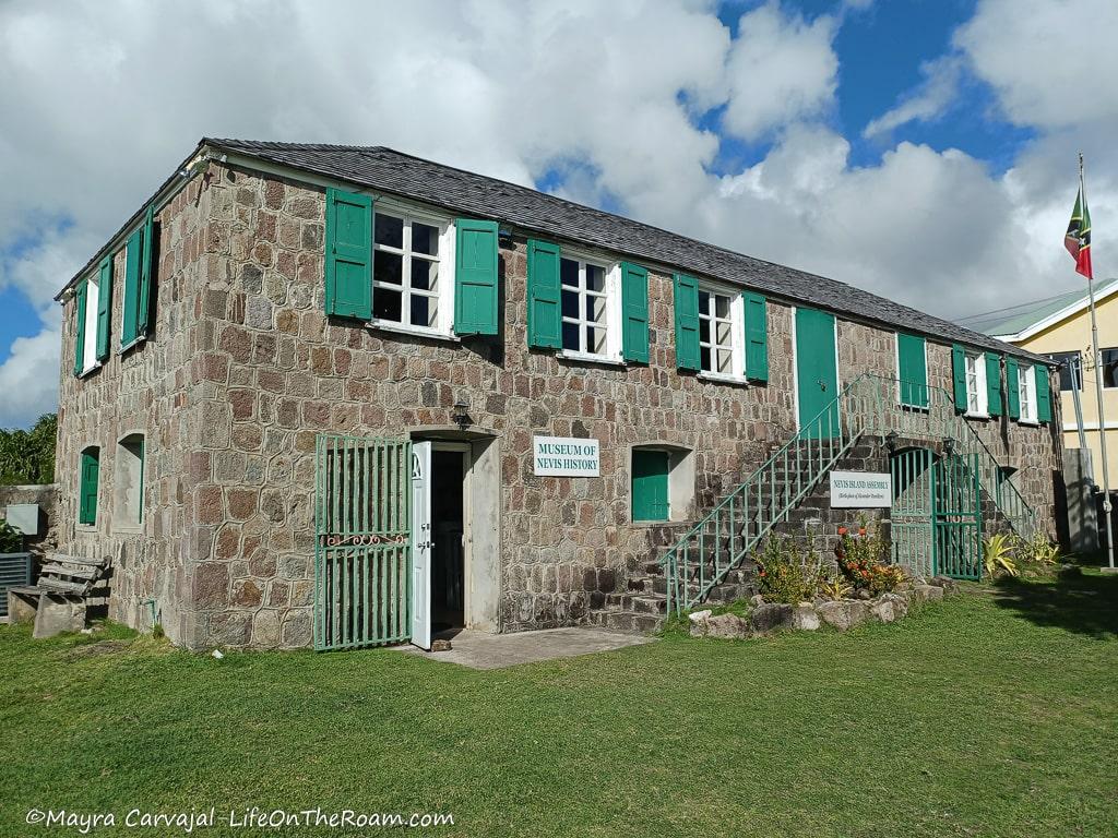 A 2-storey small stone building with turquoise hurricane shutters and the sign "Museum of Nevis History"