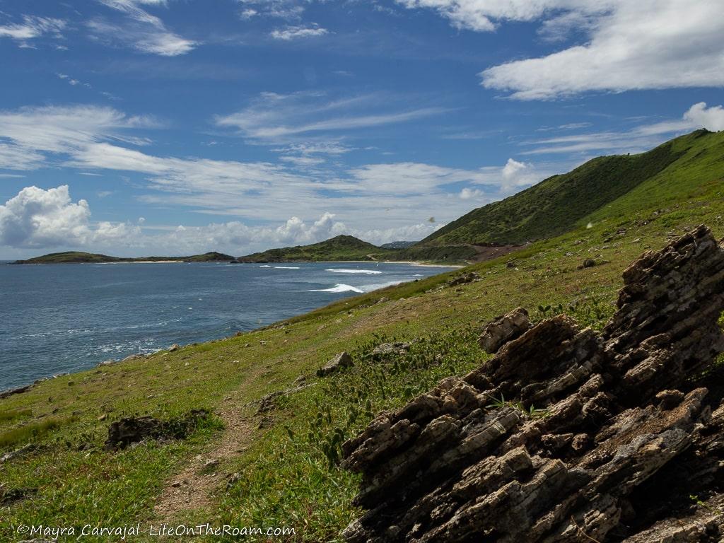 A trail along the coast with grass, rocks, and hills in the distance