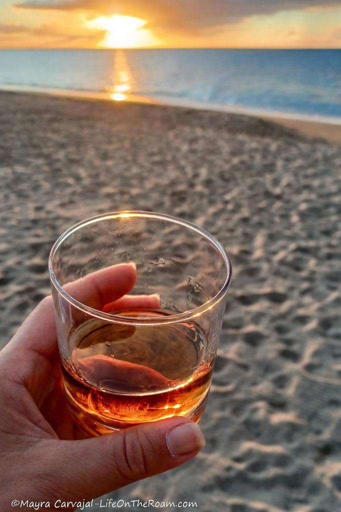 A woman holding a glass with rum on a beach at sunset
