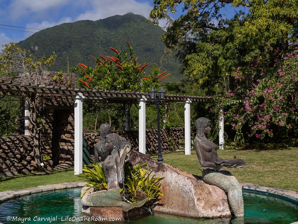 A fountain with a sculpture of three mermaids and a dormant volcano in the back