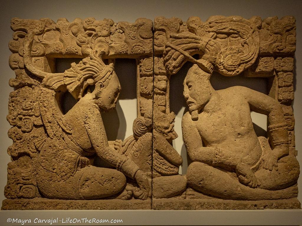 A carved stone panel with two human figures facing each other and a representation of a deity in the centre