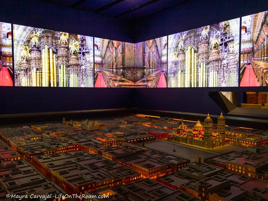 An architectural model of a city with a large screen above showing architectural details