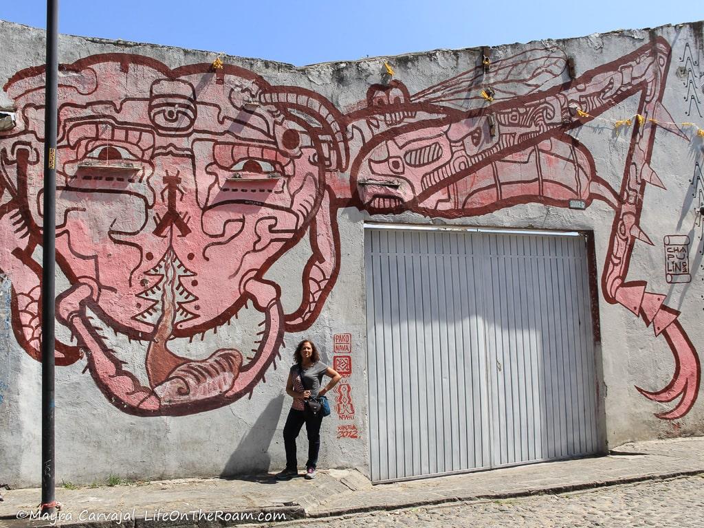 Mayra standing in front of a mural representing a grasshopper