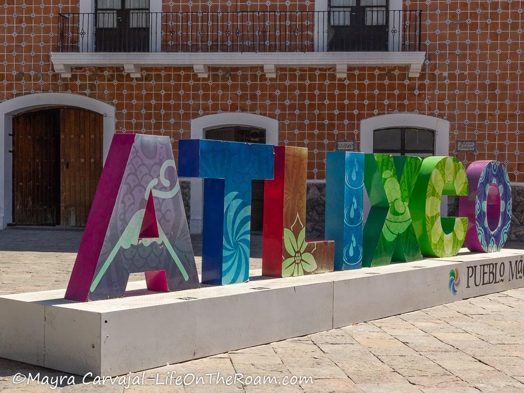 A street sign in colours with the world ATLIXCO in front of a building with tiles and brick