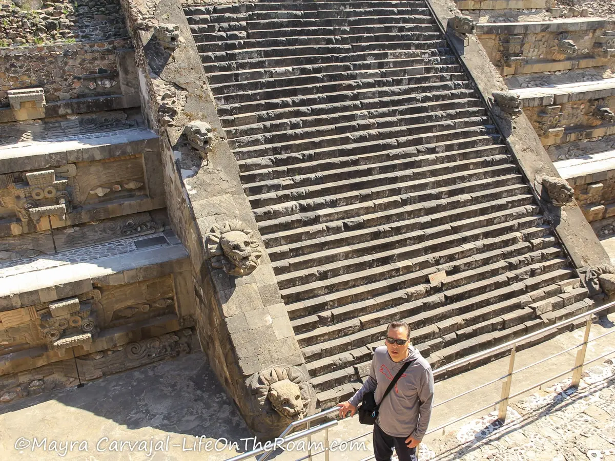 A man standing at the foot of a pyramid with a staircase adorned with stone serpent heads
