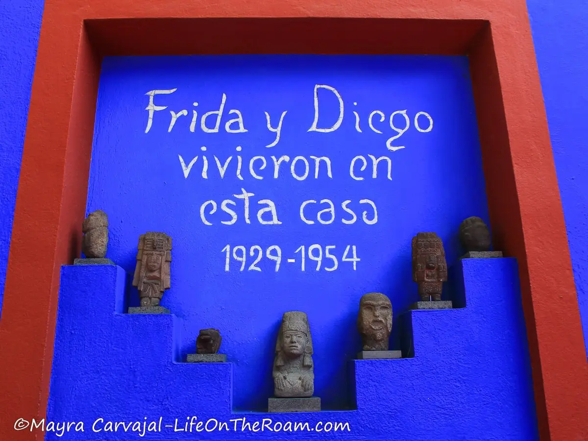A blue wall with pre-Hispanic figurines and the text "Frida y Diego vivieron en esta casa"