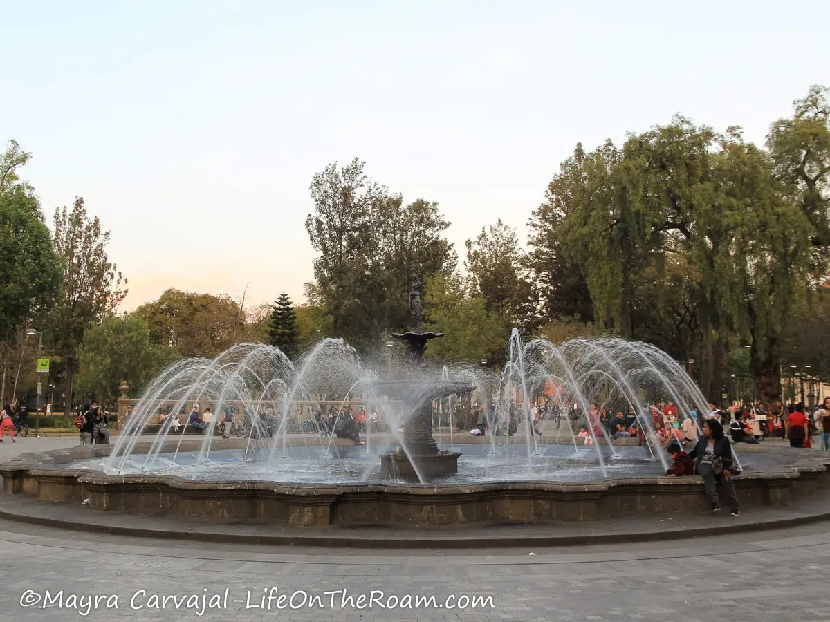 A circular fountain with a statue in the centre, in a park with trees.