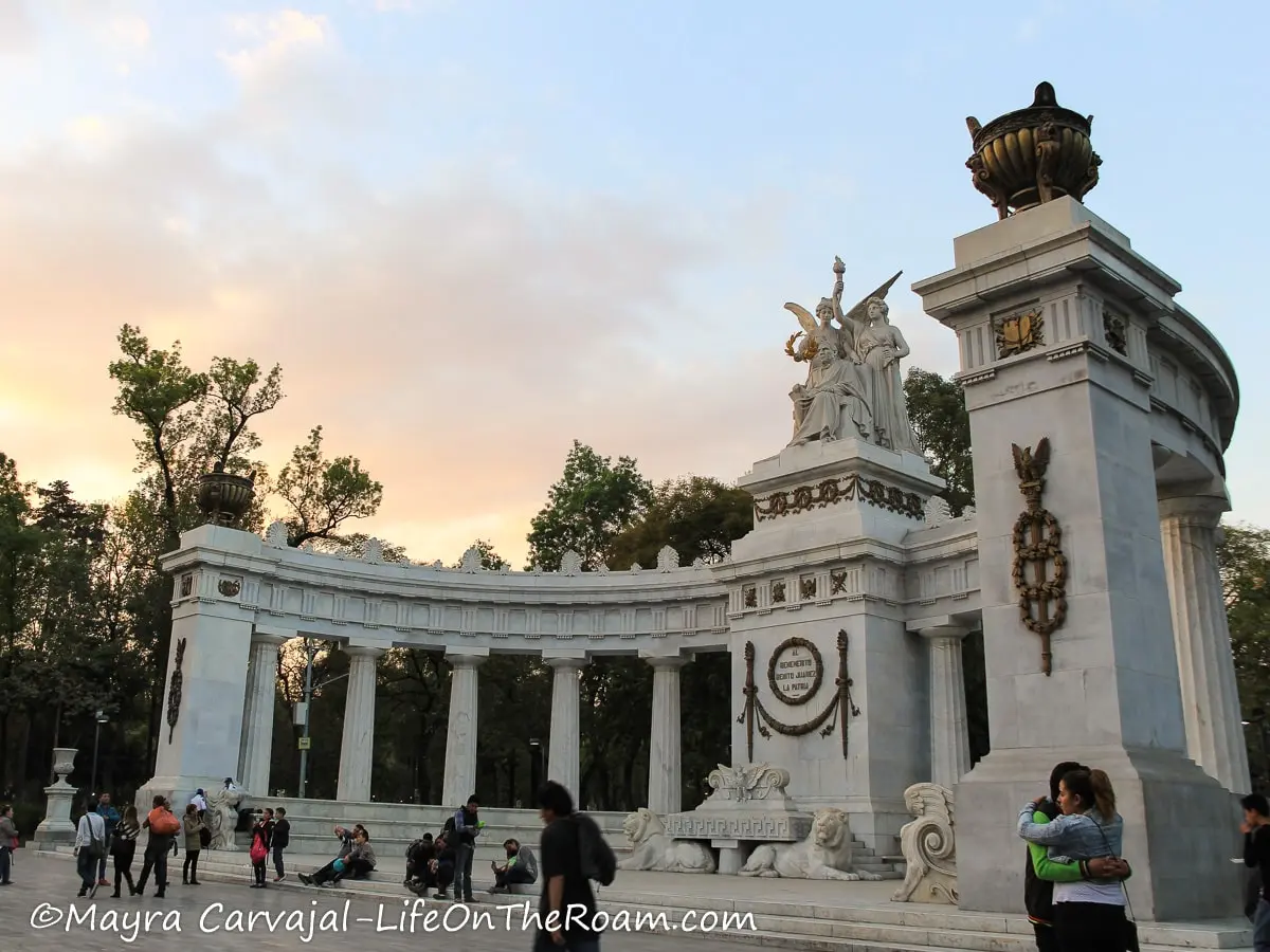 A semicircular monument in marble with columns and statues, inside a park