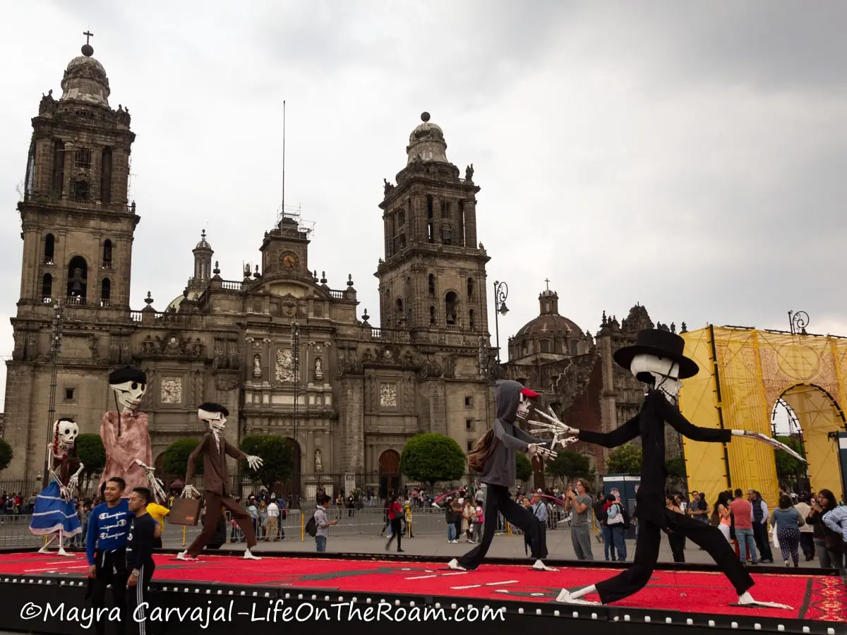 A Day of the Dead display with monumental dressed up skeletons in a square with a cathedral in the background