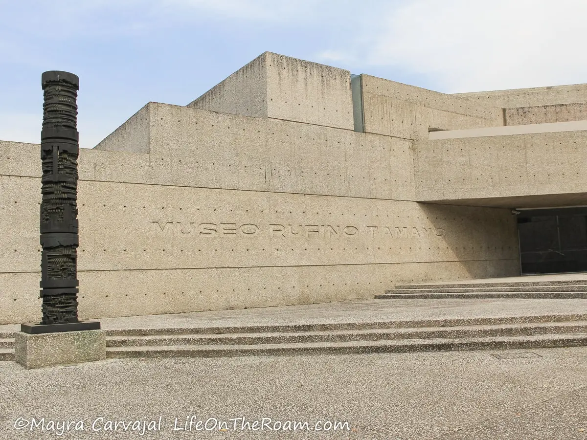 A building with bare concrete walls with the words "Museo Rufino Tamayo", and a sculpture on the left
