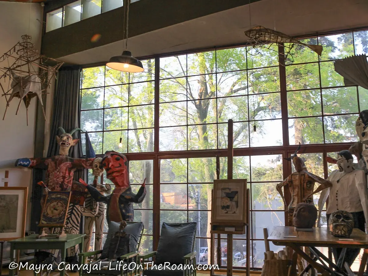 A collection of big papier mache fantastical figures and traditional toys hanging from the ceiling, skeletons on chairs, in front of a giant window