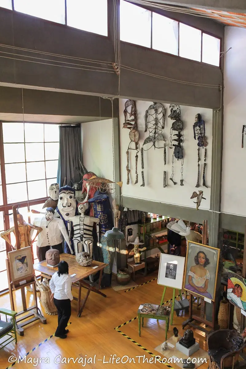 An artist's studio (Diego Rivera) with a double height ceiling, large windows, skeletons on the walls, easels, paintings, and large figures made of papier maché.