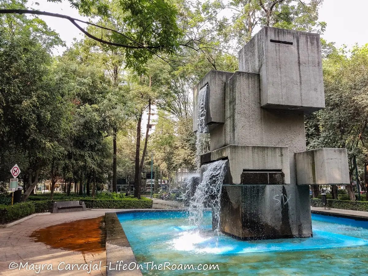 A concrete fountain with solid square shapes, in an urban park with trees
