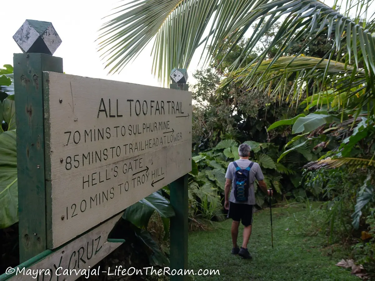 A man entering a trail in a tropical environment with a sign saying "All Too Far Trail"