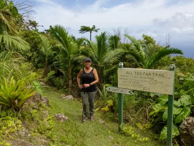 Mayra standing on a trail with grass on the path and tall palm trees along the edge, the sea in the background and a sign with names of trails on Saba