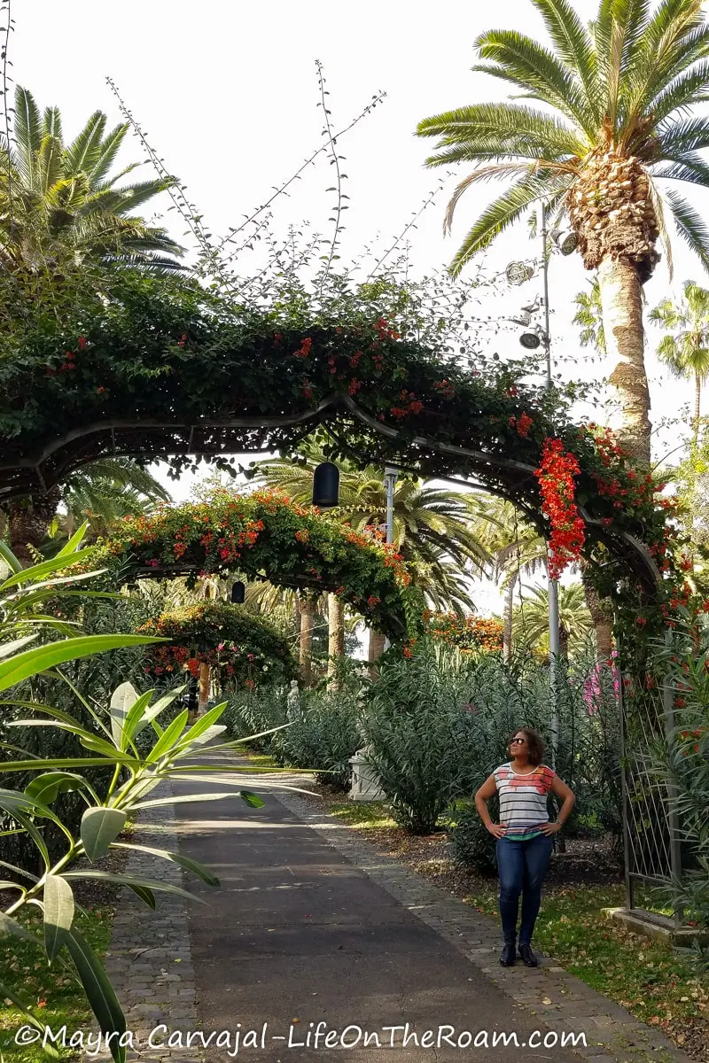 Mayra standing at the entrance of a series of arches with flowers and leaves with palm trees in the background