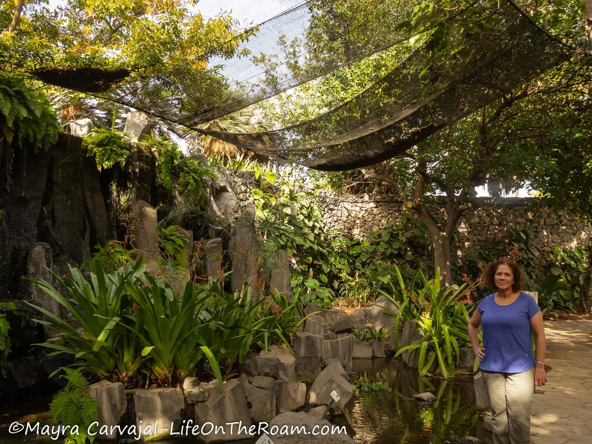 Mayra standing under a canopy in a shaded garden with a waterfall and tropical vegetation