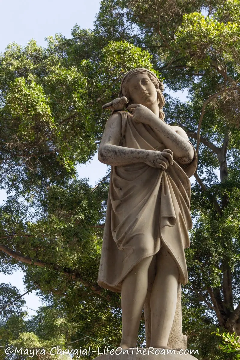 Stone sculpture of a muse-like human figure with a bird on the shoulder, with trees against the sky in the background