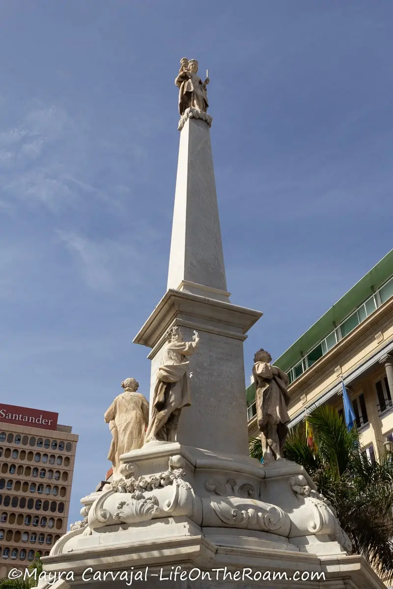 A big monument with an obelisk showing a Virgin at the top and four figures at the base of the monument