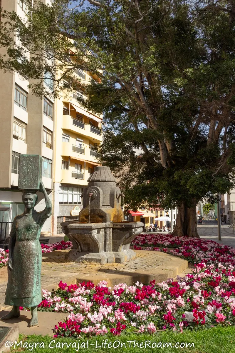 A square with an outer circle of flowers, a small fountain in the centre, and a sculpture of a woman in the forefront