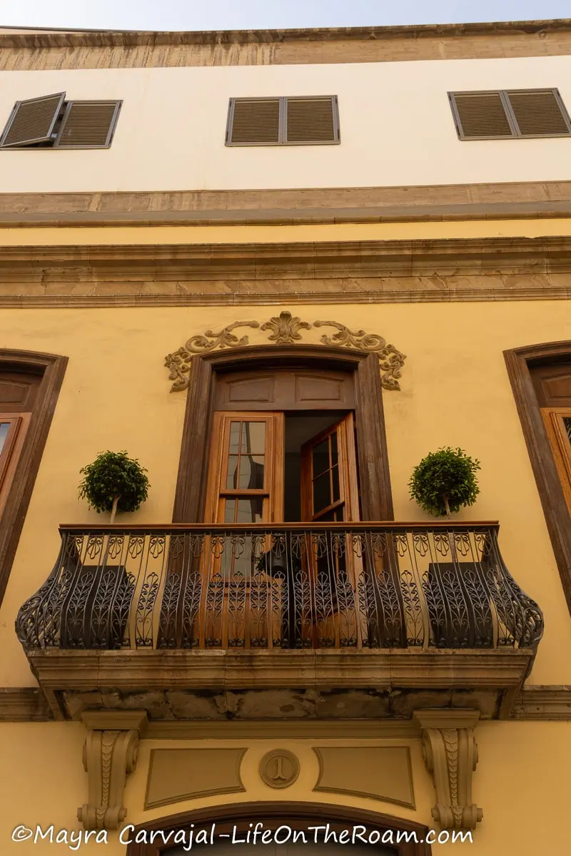A wrought-iron balcony in a muted yellow building with a decorated wooden door
