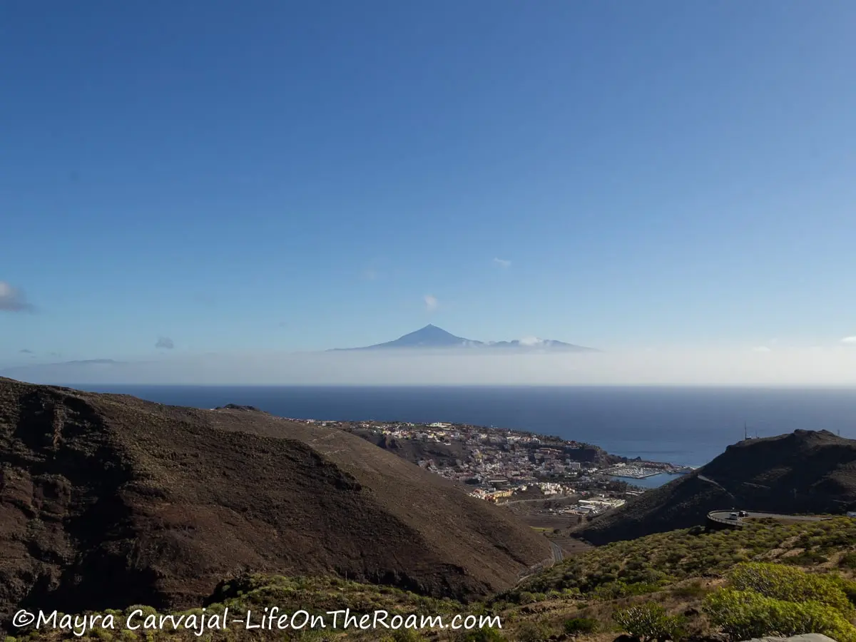 A view through the mountains of a seaside town with a volcano peak in the far distance