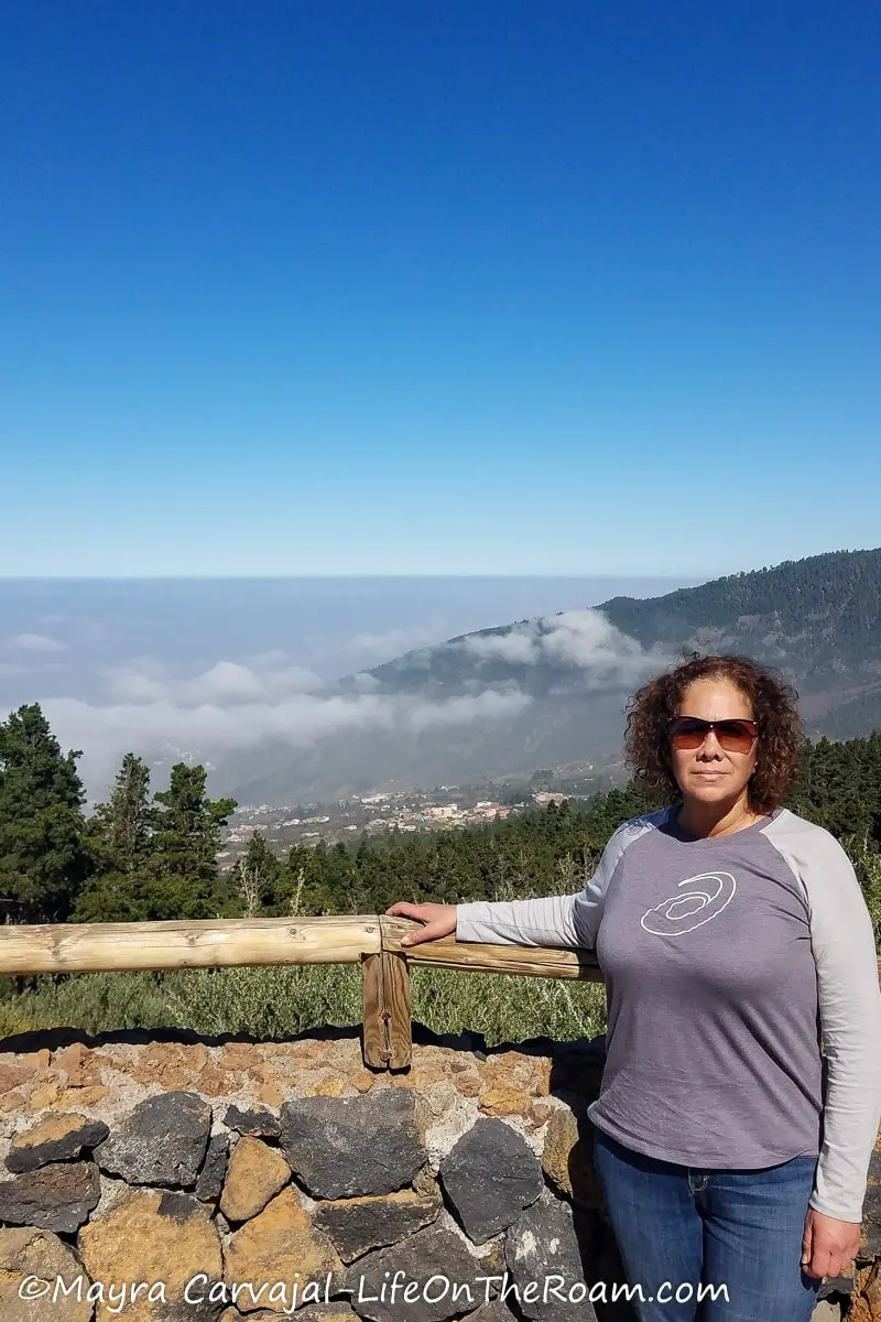 Mayra standing in a lookout overviewing a pine-covered mountain with the sea in the distance