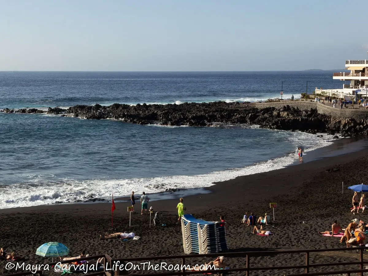 A black sand beach in a bay with sun beds and a few people on the sand