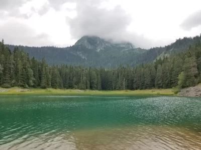 A lake with a dense pine forest around the edge and a mountain peak in the background on a cloudy day