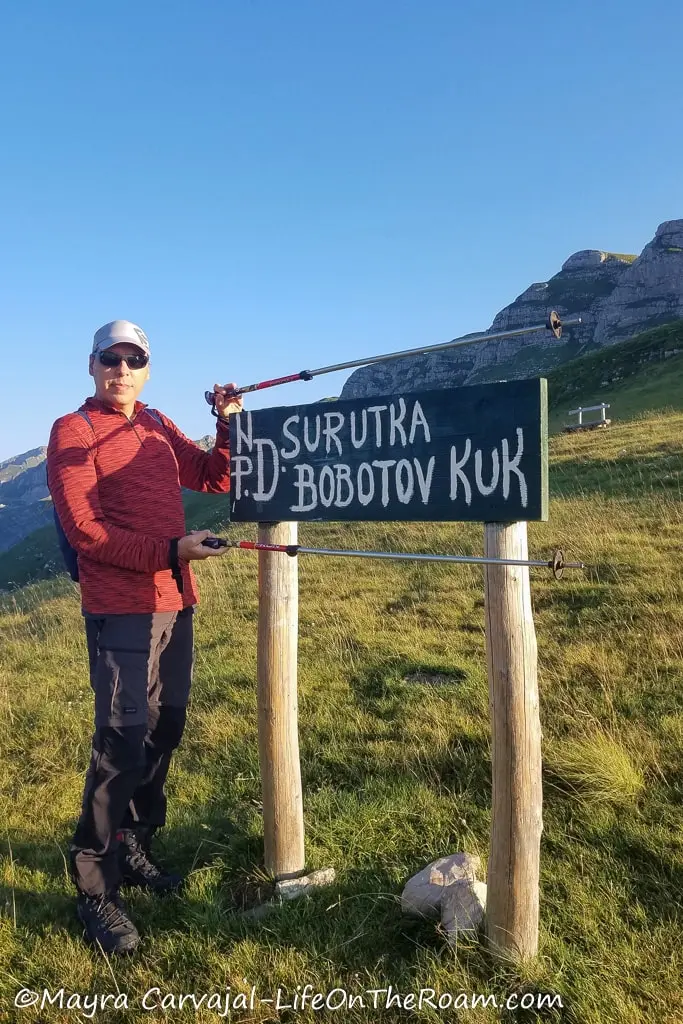 A man framing a trailhead sign with his hiking sticks, that says "Bobotov Kuk"