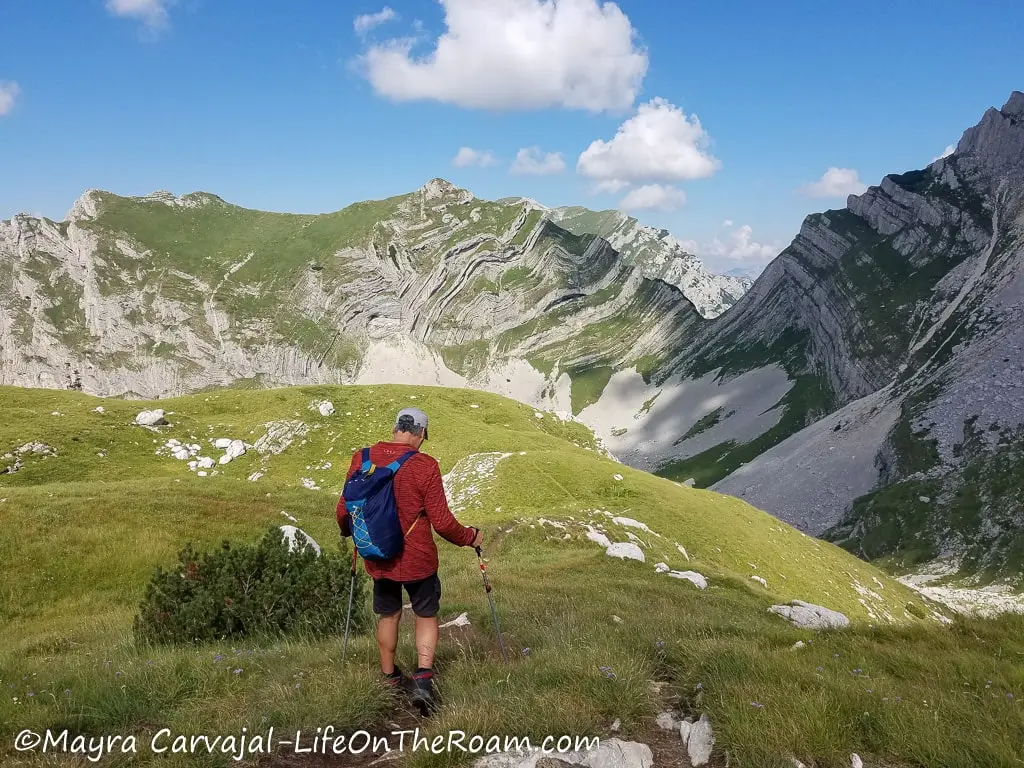 A man walking on a trail through a valley covered with grass, with a mountain showing layers in the background