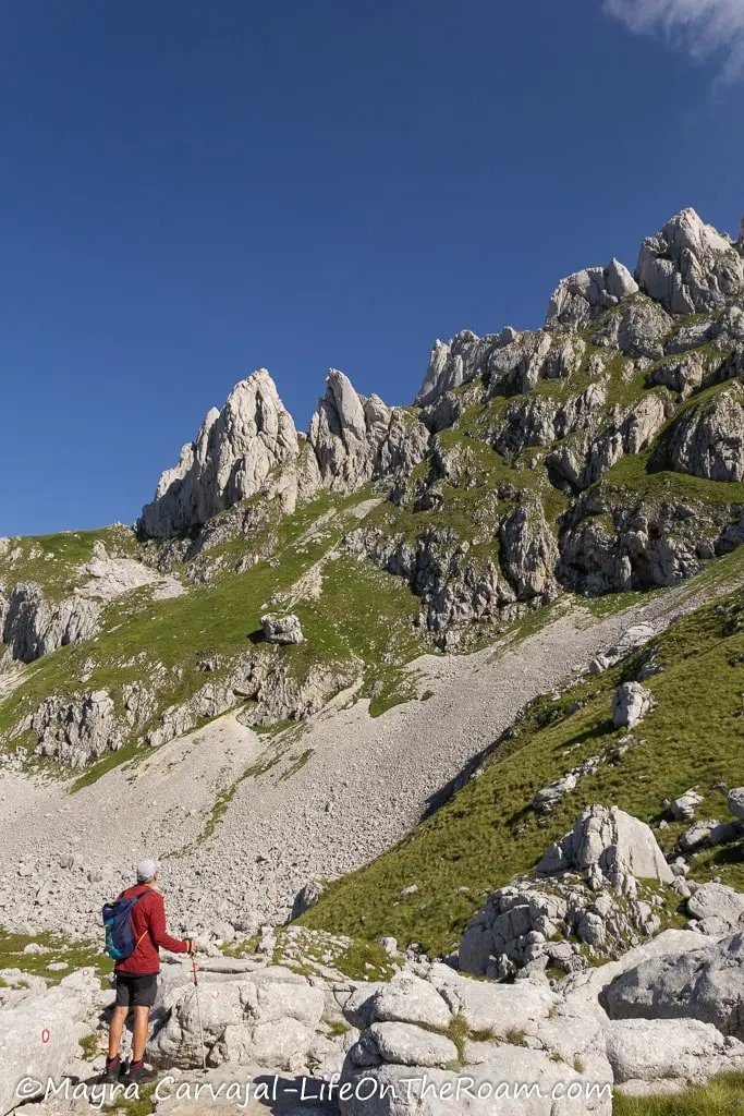 A man standing on a trail at the bottom of huge rock formations and a slope with scree