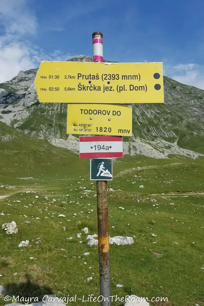 A metal post along a mountain trail with yellow signs indicating name of trails, distance, and a colour dot