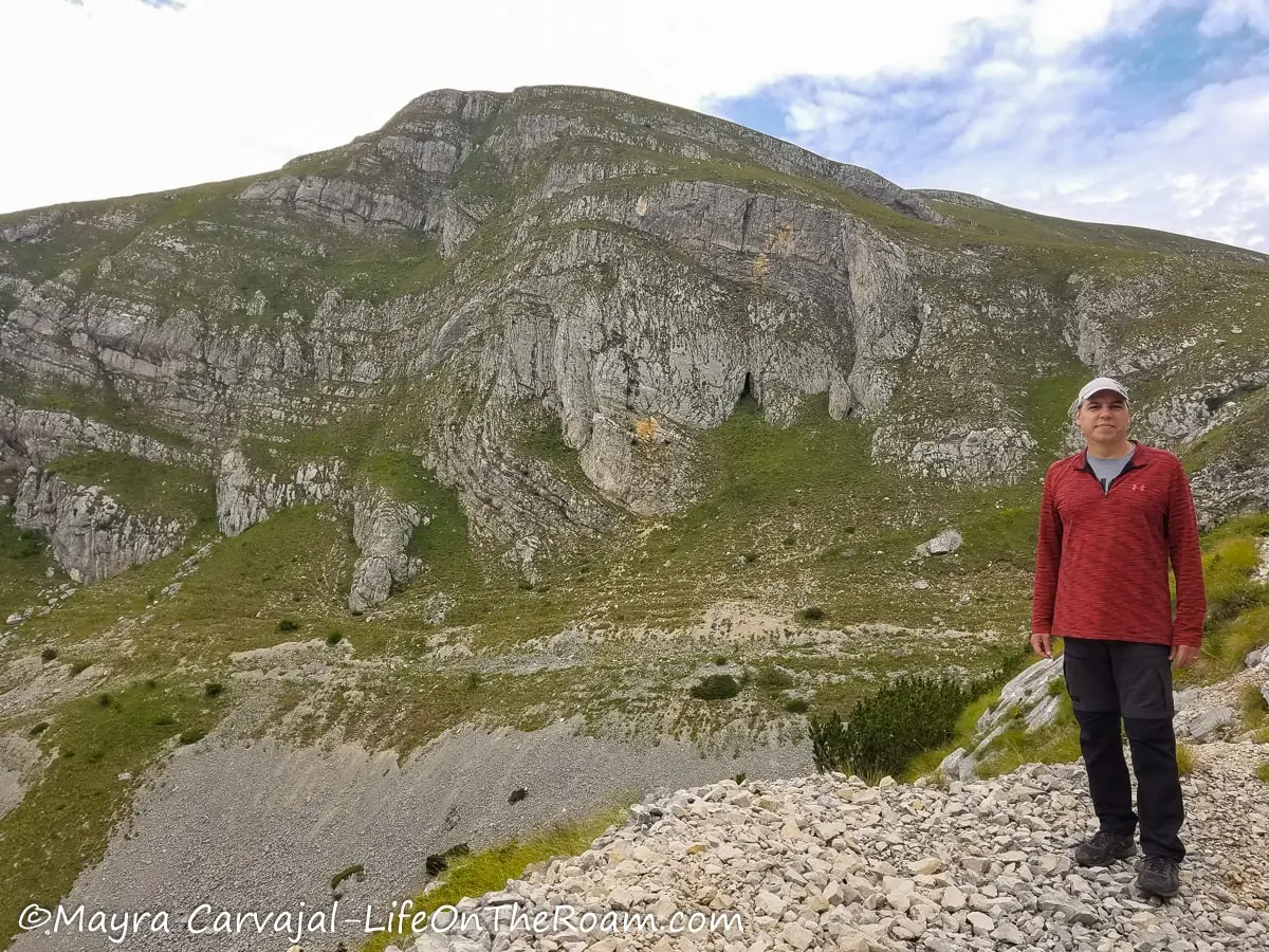 A man standing on the side of the road showing a mountain with horizontal convoluted layers