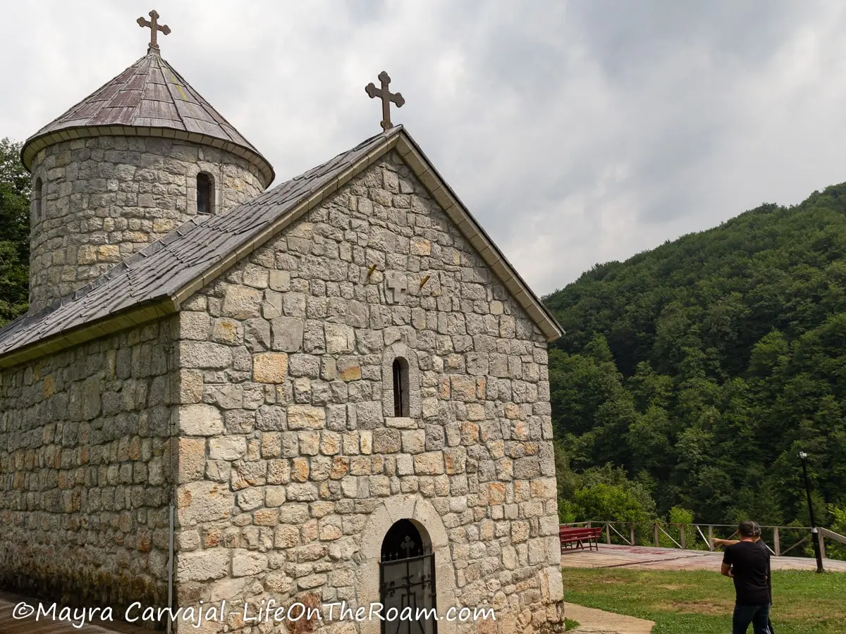 A small monastery building with a gray stone exterior, a small door and window in the front and trees in the background