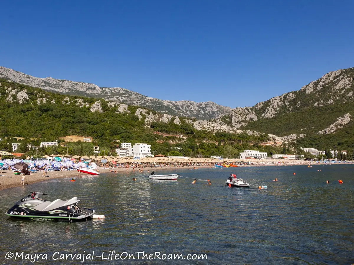 A long beach with some boats in the water and with some buildings at the foot of the hill in the background