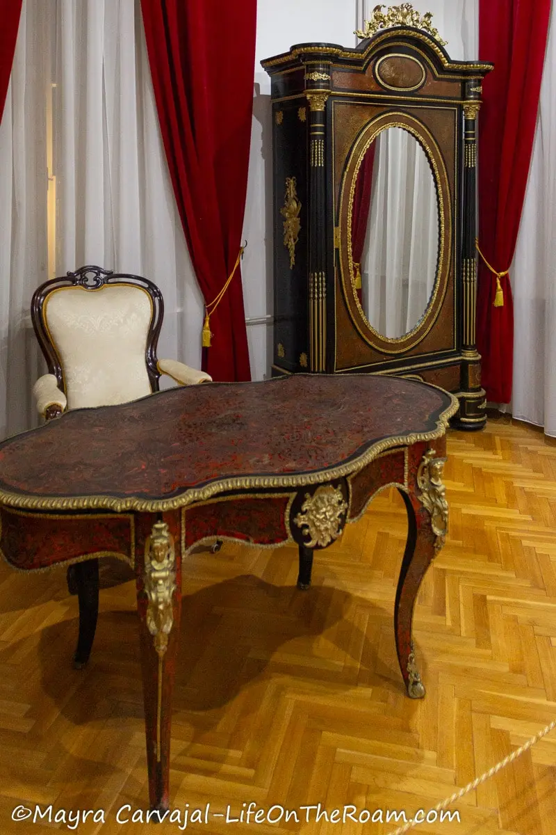 A wood desk, chair, and armoire with a mirror in classical style  with gold plated details in an exhibit setting