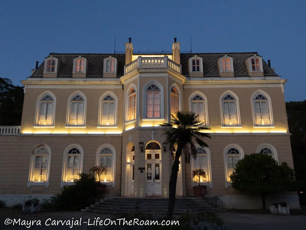 A 2-storey big residential  in palatial style of the19th century at dusk with the lights on. Building has arched windows and dormer windows at the top. 