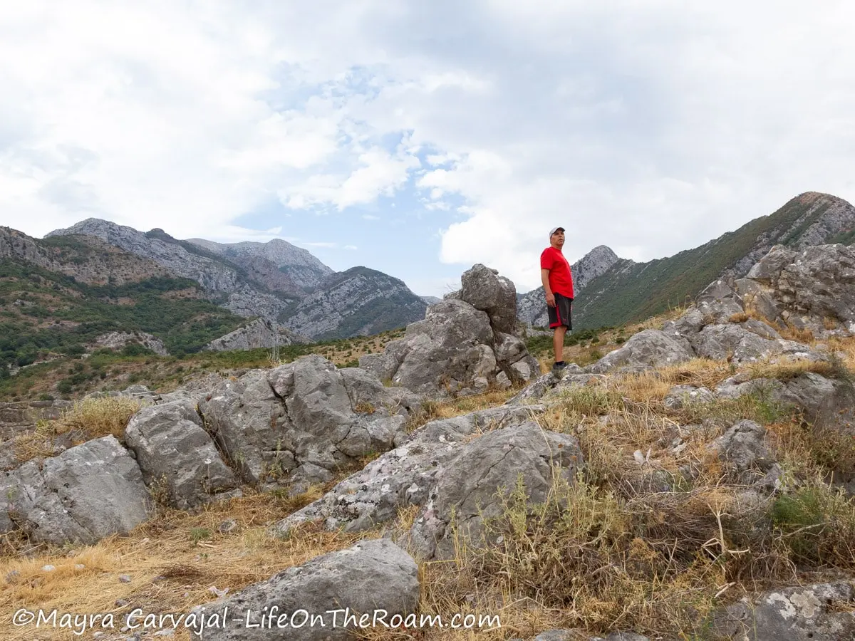 A man standing on rocks partially covered by tall grasses with mountains in the background