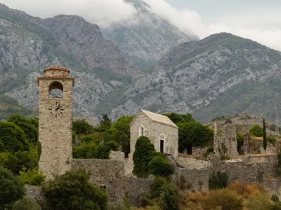 View of an ancient city in the mountains behind stone walls, with a clock tower and several old buildings in cut stone, with trees around.