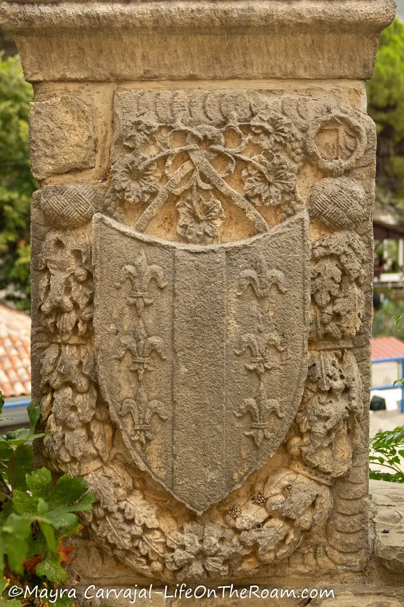 A coat of arms in stone decorated with fleur de lis and a wreath depicting leaves