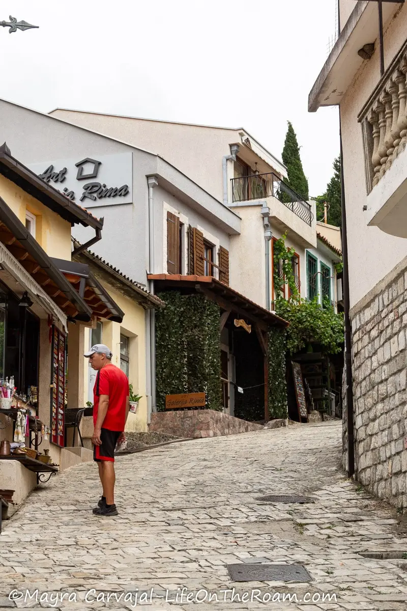 A man standing on a cobblestone street going uphill and looking at souvenirs on a shop inside 2-story houses